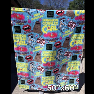 May include: A light blue blanket with a repeating pattern of a dinosaur, a cat, and text that reads "Dungeon Crawler Carl". The blanket measures 50" x 60".