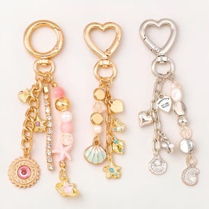 Op de afbeelding: Drie metalen sleutelhangers met diverse bedels. De eerste heeft een ronde sluiting, roze kralen, een zeester en een zon. De tweede heeft een hartvormige sluiting, schelpbedels en kralen. De derde is zilverkleurig met hart- en schelpbedels.
