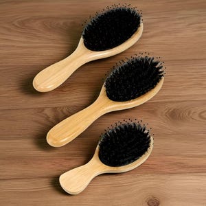 以下が含まれることがあります： 木製のヘアブラシが3つ、黒い毛で木製の表面に配置されています。ブラシは明るい茶色の楕円形のヘッドと湾曲したハンドルを持っています。毛は密に詰まっており、柔らかく見えます。