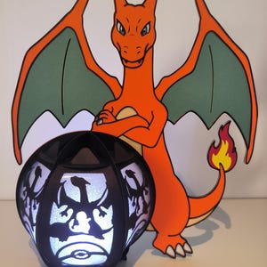 May include: A black and white 3D printed lamp with a silhouette of a dragon. The lamp is lit up and has a white glow. A red and green cartoon dragon stands behind the lamp.