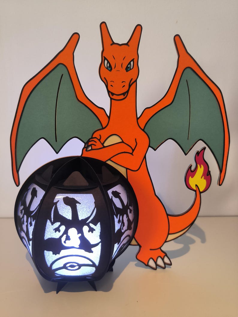 May include: A black and white 3D printed lamp with a silhouette of a dragon. The lamp is lit up and has a white glow. A red and green cartoon dragon stands behind the lamp.