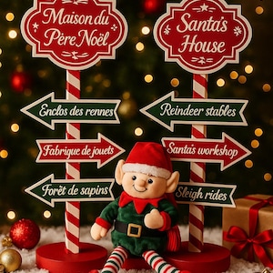 Puede incluir: Una escena navideña festiva con letreros que señalan la Casa de Santa y otros destinos. Una muñeca elfo de peluche está sentada frente a los letreros. Los letreros son rojos y blancos con flechas verdes. También se ven adornos navideños y un regalo envuelto.