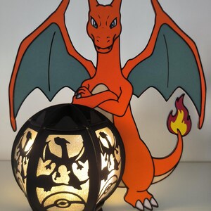 May include: A black and orange Pokemon lamp with a silhouette of Charizard on the lampshade. The lamp is lit and the light shines through the silhouette. The lamp is sitting on a white surface.