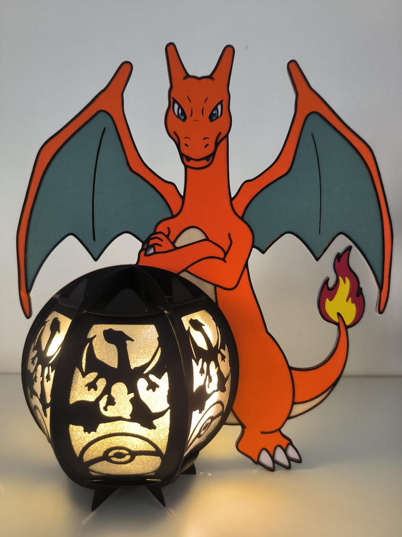 May include: A black and orange Pokemon lamp with a silhouette of Charizard on the lampshade. The lamp is lit and the light shines through the silhouette. The lamp is sitting on a white surface.