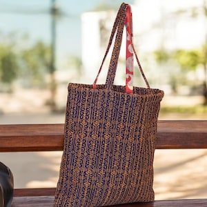 May include: A blue and gold patterned tote bag with red and white fabric straps. The bag is sitting on a wooden bench.