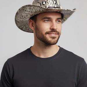 Op de afbeelding: Cowboyhoed met een slangenprint in grijstinten en beige. De hoed heeft een brede rand en een zwarte band. De hoed wordt gedragen door een persoon die een zwart t-shirt draagt.