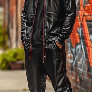 May include: A person wearing a black leather jacket with red trim, matching pants, and patterned high-top sneakers. The outfit includes a black t-shirt and sunglasses. The background features a brick wall with graffiti.