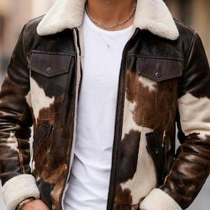 May include: A brown and white cowhide-patterned leather jacket with a cream-colored shearling collar and cuffs. The jacket has two front pockets and a zipper closure. The person is wearing a white t-shirt and black jeans.