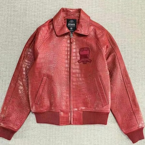 May include: A red leather jacket with a crocodile skin texture. The jacket has a collared neckline, a zipper closure, and a patch on the chest. The jacket has ribbed cuffs and hem. The brand name "AVIREX" is visible on a tag.