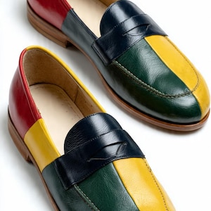 May include: A pair of loafers featuring a multi-colored design with panels of red, yellow, dark green, and navy blue leather. The shoes have a classic penny loafer strap across the vamp, a rounded toe, and a low brown stacked heel and sole.