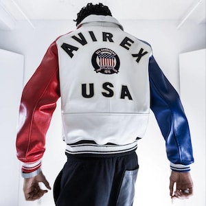 Puede incluir: Chaqueta de cuero blanca con mangas rojas y azules. La parte trasera de la chaqueta presenta la palabra "AVIREX" sobre un emblema circular con las palabras "ALL AMERICANS" y "USA" debajo. La chaqueta tiene cuello y puños acanalados.