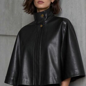 May include: Black leather cape with a high collar and two gold buttons. The cape has short sleeves and a tailored fit. The model is wearing black pants. The background is a neutral gray wall.