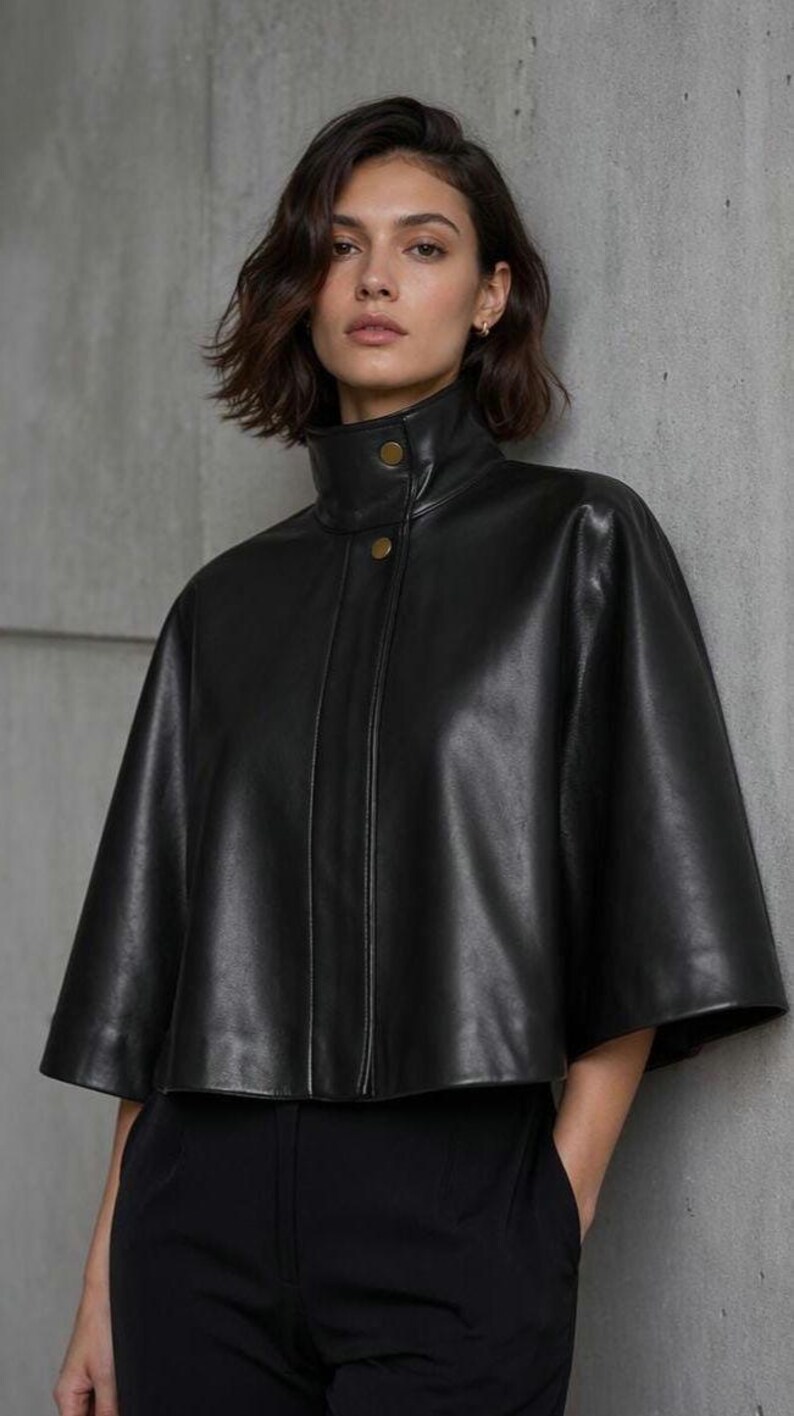 May include: Black leather cape with a high collar and two gold buttons. The cape has short sleeves and a tailored fit. The model is wearing black pants. The background is a neutral gray wall.