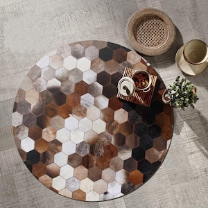 May include: A circular rug featuring a geometric design of brown, tan, and white hexagonal tiles. The rug has a natural, textured look and is positioned on a light grey wooden floor. A woven basket, a small plant, and a tray with a can and cup are also visible.
