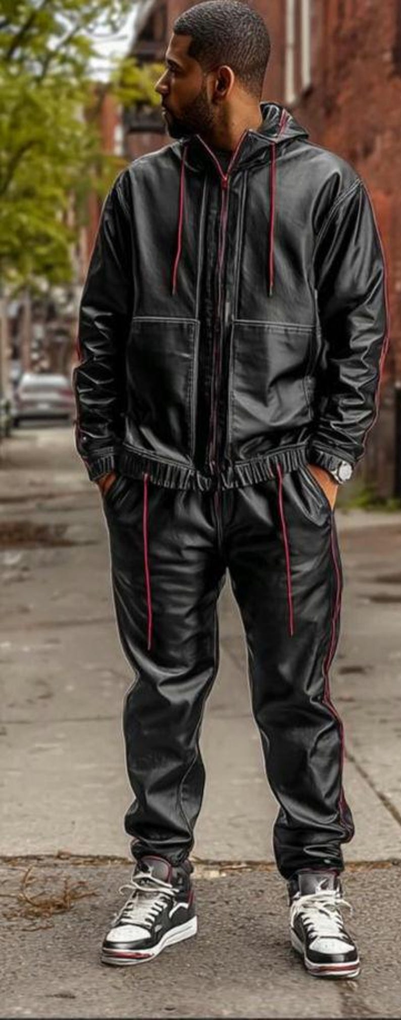 May include: Black leather tracksuit with red trim. The jacket has a hood and a zip closure. The pants have a drawstring waist. The outfit is paired with black and white sneakers.
