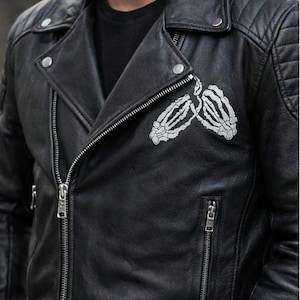 Puede incluir: Chaqueta de cuero negra de estilo motero con cremallera plateada y un diseño de manos de esqueleto blancas en el pecho. La chaqueta tiene un detalle de hombro acolchado y un estilo motero clásico.