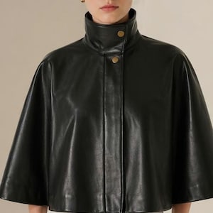 May include: A black leather cape with a high collar and two gold buttons. The cape is worn over white pants. The model is wearing gold hoop earrings. The garment has a sleek, modern design, suitable for outerwear.