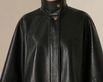 Può includere: Mantella in pelle nera con collo alto e due bottoni dorati. Il modello indossa orecchini a cerchio dorati. Il capo ha un design elegante e moderno, adatto all'abbigliamento da esterno. Il modello indossa pantaloni bianchi.