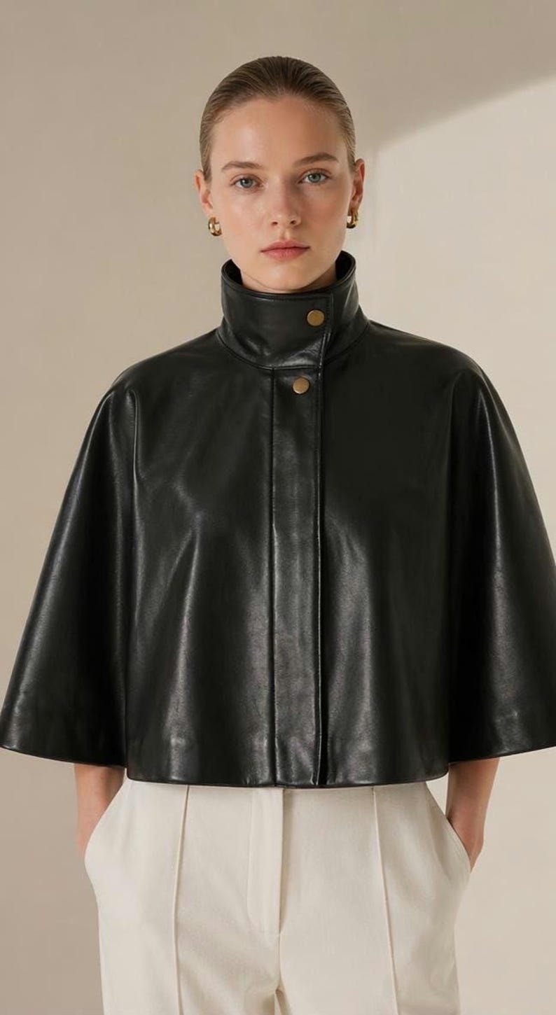 May include: A black leather cape with a high collar and two gold buttons. The cape is worn over white pants. The model is wearing gold hoop earrings. The garment has a sleek, modern design, suitable for outerwear.