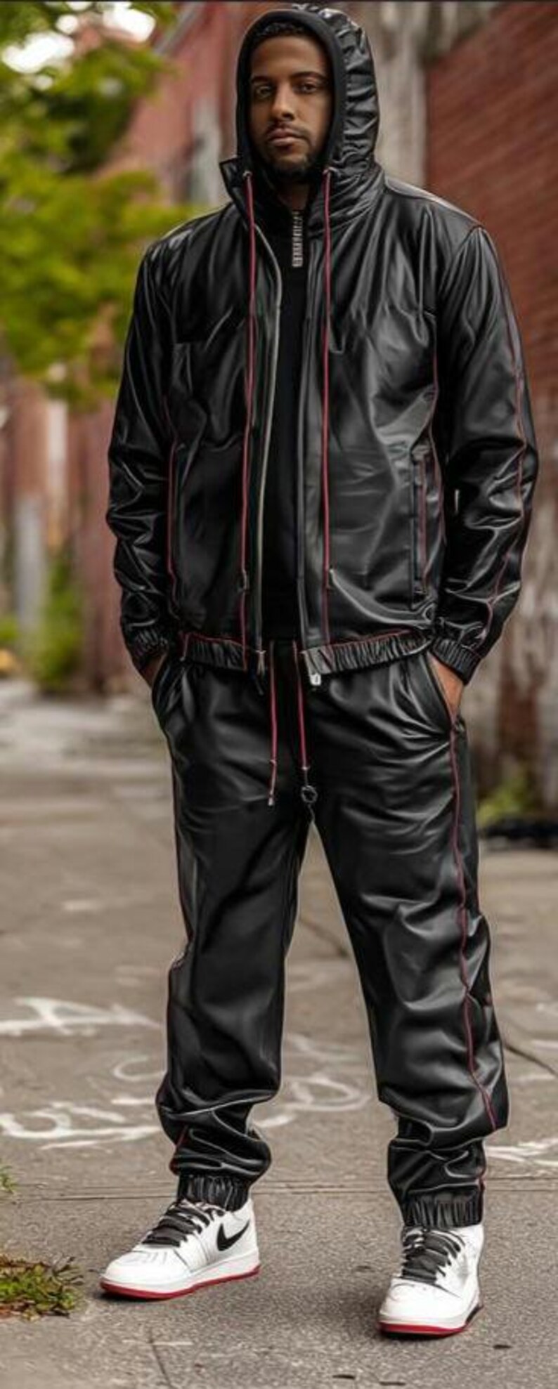 May include: Black hooded jacket and matching pants with red trim. The outfit is made of a shiny material. The person is wearing white sneakers with black laces and a red sole.