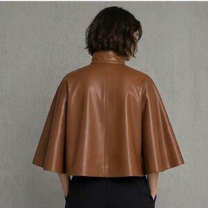 May include: A brown leather cape jacket with a high collar. The jacket has a flared, cape-like design and is made of a smooth, shiny leather material. The model is wearing black pants.
