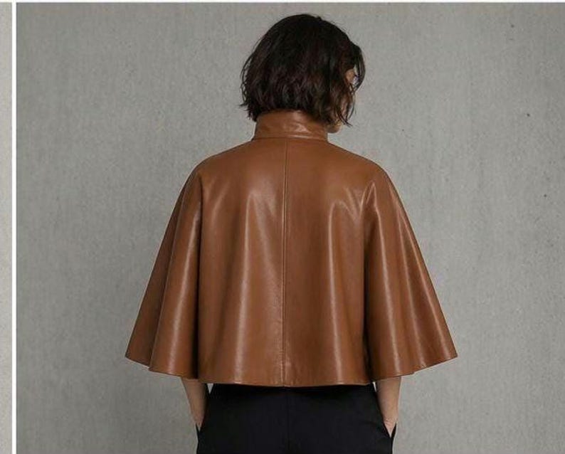 May include: A brown leather cape jacket with a high collar. The jacket has a flared, cape-like design and is made of a smooth, shiny leather material. The model is wearing black pants.