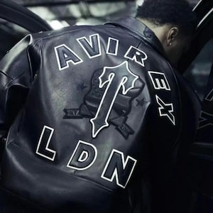Könnte beinhalten: Schwarze Lederjacke mit weißem Schriftzug "AVIREX LDN" auf dem Rücken. Ein großes Emblem mit einem "T"-Design ist unterhalb der Schrift angebracht. Die Jacke wird von einer Person getragen, die sich in ein Auto lehnt.