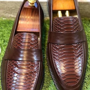 Könnte beinhalten: Ein Paar braune Leder-Loafer mit Schlangenmuster auf dem Blatt und einem glatten Lederriemen. Die Schuhe haben ein klassisches Design mit runder Spitze und stehen auf einer grünen Oberfläche.