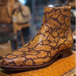 May include: A high-top boot with a snakeskin pattern in shades of yellow and brown. The boot has a pointed toe and a low heel. The boot is displayed on a wooden surface.