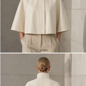 May include: Ivory-colored cape jacket with a high collar and button closure. The jacket has short, wide sleeves and is paired with matching beige trousers. The word "IVORY" is printed on the trousers. The model is wearing the jacket.
