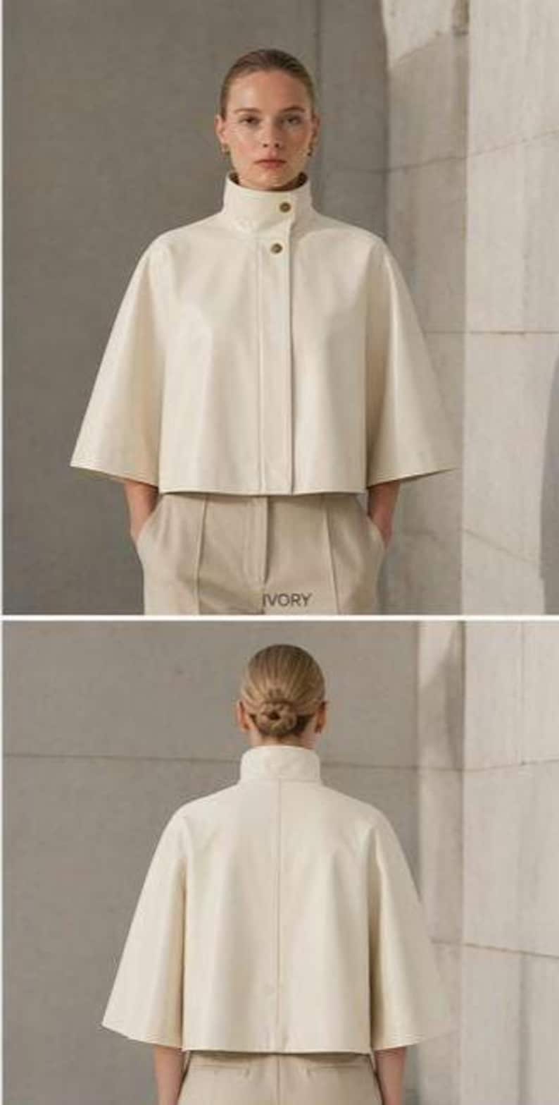 May include: Ivory-colored cape jacket with a high collar and button closure. The jacket has short, wide sleeves and is paired with matching beige trousers. The word "IVORY" is printed on the trousers. The model is wearing the jacket.
