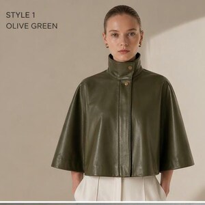 May include: Olive green leather cape with a high collar and button closure. The garment is styled with white pants. The text "STYLE 1 OLIVE GREEN" is in the upper left corner.