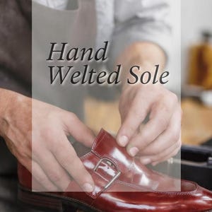 Może przedstawiać: Polerowany bordowy sk&oacute;rzany but z klamrą, srebrną metalową sprzączką i widoczną szytą podeszwą. Tekst "Hand Welted Sole" jest nałożony na obraz wyraźną, ciemną czcionką szeryfową na jasnym tle.