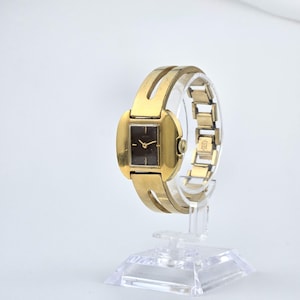 Può includere: Un orologio da polso vintage color oro con un quadrante rettangolare scuro. L'orologio presenta un cinturino dorato ed è esposto su un supporto trasparente. L'orologio ha un design classico, che mostra un'estetica senza tempo.