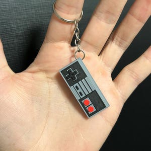 Pode incluir: Um chaveiro de controlador Nintendo Entertainment System (NES) cinza e preto com botões vermelhos. O chaveiro está preso a um chaveiro prateado.