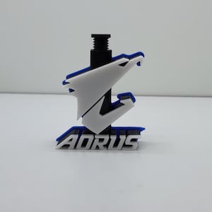 Pode incluir: Um logotipo AORUS impresso em 3D em branco, azul e preto. O logotipo está montado em um parafuso preto. O logotipo é uma águia estilizada com as asas abertas.