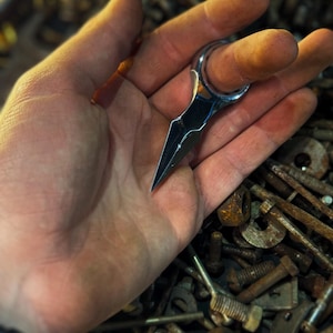 Könnte beinhalten: Ein kleines, silberfarbenes Ringmesser mit einer spitzen Klinge. Das Messer wird in einer Hand gehalten, wobei der Ring um einen Finger liegt. Der Hintergrund ist ein Haufen rostiger Metallbeschläge.