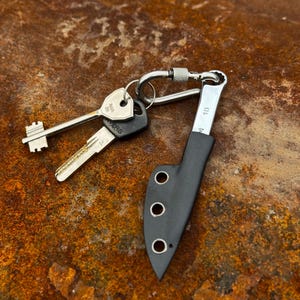 May include: A small, black knife in a sheath attached to a keyring with two keys. The knife has a silver blade and a black handle. The keyring is silver and has a carabiner clip. The keys are silver with black plastic key heads.