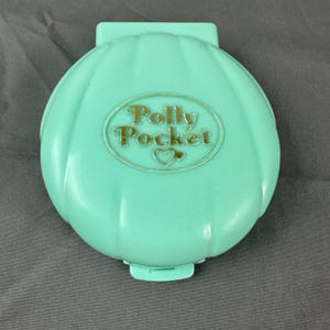 Könnte beinhalten: Ein mintgrünes Kunststoffgehäuse in Muschelform mit Klappdeckel. In der Mitte befindet sich ein ovales, geprägtes Logo mit dem Schriftzug Polly Pocket und einem kleinen Herzsymbol. Die Außenfläche weist vertikale Rillen wie eine Muschel auf.