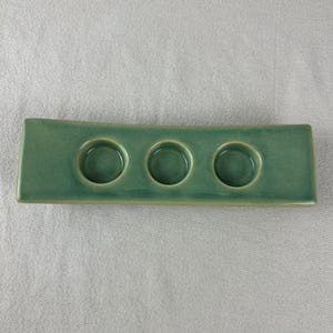 May include: A rectangular, green ceramic candle holder with three circular indentations. The holder has a slightly curved shape and a glossy finish. The color is a muted, earthy green, suitable for home decor.