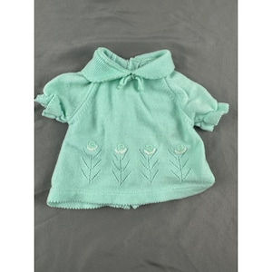 Könnte beinhalten: Hellgrüner Baby-Pullover mit Bubikragen und kurzen Ärmeln. Der Pullover hat eine Reihe gestickter weißer Blumen. Der Kragen hat eine kleine Schleife. Der Pullover ist aus weichem Strickmaterial.