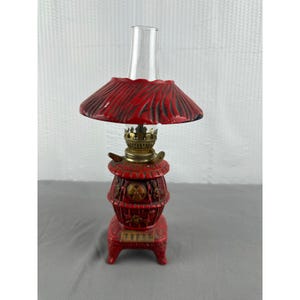 May include: A vintage red oil lamp with a glass chimney. The lamp has a decorative base and a red shade. The lamp's metal components are gold-colored. The lamp is a decorative item.