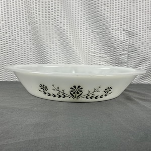 Peut inclure: Un plat de cuisson ovale blanc en verre opalin orné d'un motif floral noir imprimé sur le côté. Le design se compose d'une fleur centrale rappelant une marguerite, entourée de petites fleurs et de tiges feuillues s'étendant horizontalement.