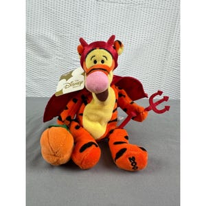 Könnte beinhalten: Ein Plüschtier von Tigger als Teufel für Halloween. Der orange und schwarz gestreifte Tigger trägt ein rotes Teufelskostüm mit Dreizack und Kürbis. Das Disney Store-Etikett ist sichtbar.