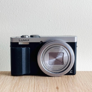 Op de afbeelding: Een zwart-zilveren Lumix digitale camera met een Leica-lens en een metalen ring. De camerabehuizing heeft een zwarte, getextureerde grip aan de linkerkant, een zilveren bovenplaat met bedieningsknoppen en de tekst Lumix en Leica op de voorzijde.
