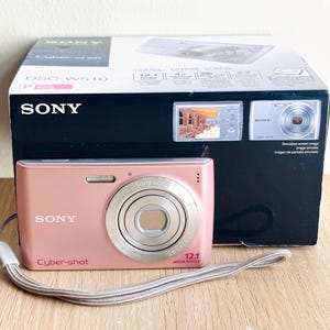 May include: A pink Sony Cyber-shot DSC-W510 digital camera with a silver lens and a grey wrist strap, positioned in front of its original product packaging box which displays the camera model, brand name, and technical specifications.
