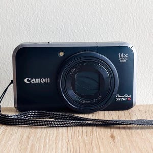 以下が含まれることがあります： シルバーの縁取りが施された黒いCanon PowerShot SX210 ISデジタルカメラ。前面には5.0-70.0mm 1:3.1-5.9の仕様が記されたレンズ、14x optical zoomのラベルがあり、カメラ本体の側面には黒いリストストラップが取り付けられています。