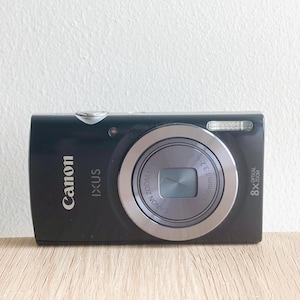 Op de afbeelding: Een zwarte Canon IXUS digitale camera met een zilverkleurige lensring, 8x optische zoomlens en ingebouwde flitser. De voorzijde toont de merknamen Canon en IXUS in witte tekst, samen met de technische specificaties van de lens.