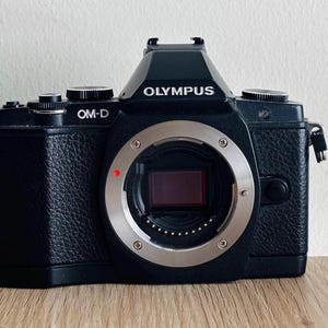 Può includere: Fotocamera digitale Olympus OM-D nera con attacco obiettivo argentato. Il corpo macchina ha una finitura strutturata e presenta il marchio "OLYMPUS" e il modello "OM-D". L'attacco dell'obiettivo ha un punto rosso.