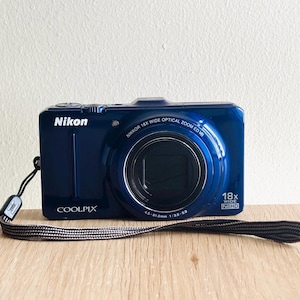 Pode incluir: Uma câmara digital Nikon Coolpix azul-escura com uma lente Nikkor 18x wide optical zoom ED VR com especificações de 4.5-81mm 1:3.5-5.9. O corpo da câmara inclui uma correia de pulso preta e exibe o texto Nikon, Coolpix e 18x Wide Full HD.